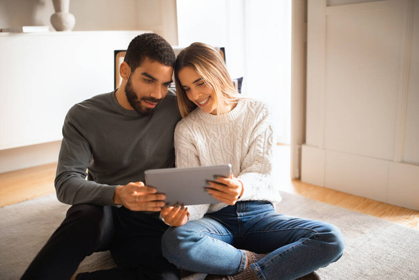 Glad millennial european wife shows tablet to arab husband, watch video, sit on floor in room interior. Love, relationships, app for blog and social network, ad and offer, video call remote at home - Photo, Image