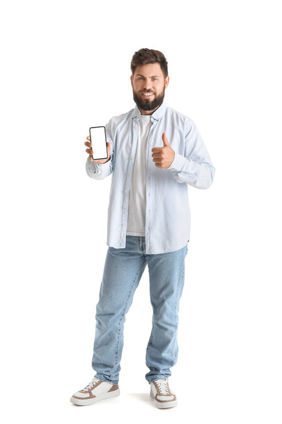 Young bearded man with mobile phone showing thumb-up on white background - 写真・画像
