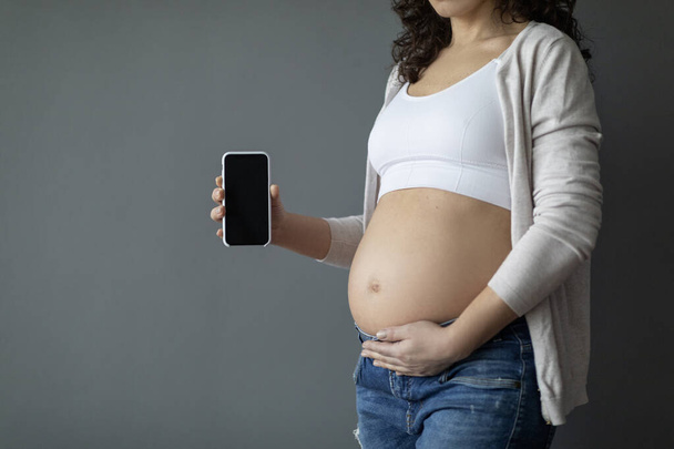 Unrecognizable Pregnant Woman Embracing Belly And Showing Blank Smartphone With Black Screen, Expectant Female Advertising Pregnancy App, Demonstrating Cellphone With Empty Screen, Mockup, Cropped - Foto, imagen