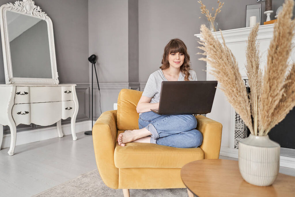 Mooi vrolijk meisje met behulp van zwarte laptop terwijl zitten op de bank in de woonkamer thuis. Jonge casual vrouw die laptop gebruikt voor communicatie en werk. S - Foto, afbeelding