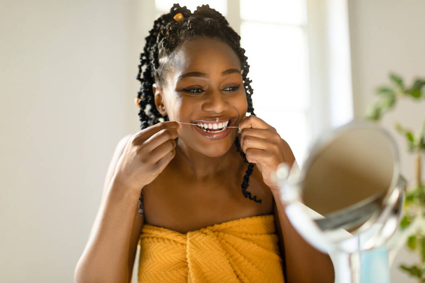 Attractive black woman looking at mirror and using dental floss, cleaning her white teeth, sitting at vanity table in bedroom interior, copy space. Dental care concept - Фото, изображение