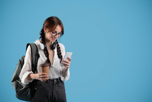 Smiling teen european girl pupil with pigtails, backpack in glasses, headphones typing on phone, drink cup of coffee takeaway on blue studio background. Study and knowledge at school, app for chat - Φωτογραφία, εικόνα