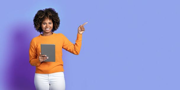 Vidám, göndör fiatal fekete hölgy alkalmi modern digitális pad a kezében mutat másolás helyet hirdetés felett lila stúdió háttér, mosolygós kamera, banner - Fotó, kép
