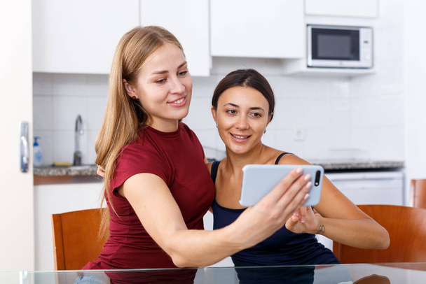 Smiling young female friends taking selfie by phone in home interior - Foto, Imagen