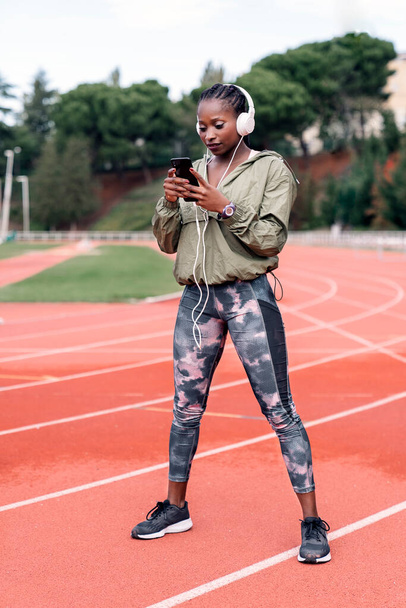 Stock photo of an African-American sprinter sitting on the athletics track with her helmets on, listening to music with her mobile phone - Foto, afbeelding