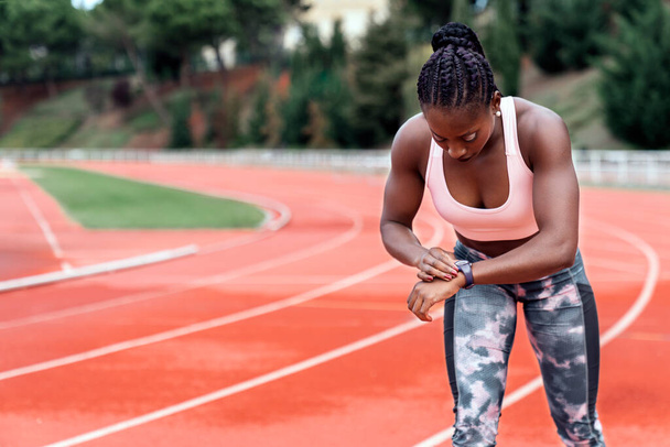 Stock photo of an African-American sprinter standing on an athletics track looking at her watch ready to start running and her hair tied up - Zdjęcie, obraz