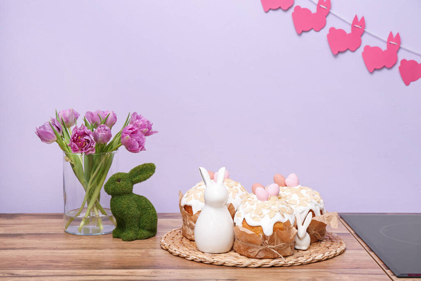 Bolos de Páscoa, coelhos e vaso com flores de tulipa no balcão da cozinha perto da parede lilás - Foto, Imagem