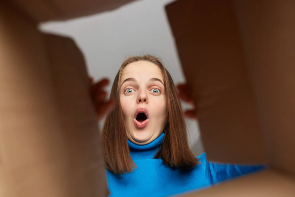 Shocked amazed Caucasian woman customer with brown hair wearing blue turtleneck opening parcel, looking inside cardboard box with shocked face, saying wow, unexpected package. - Foto, Imagem