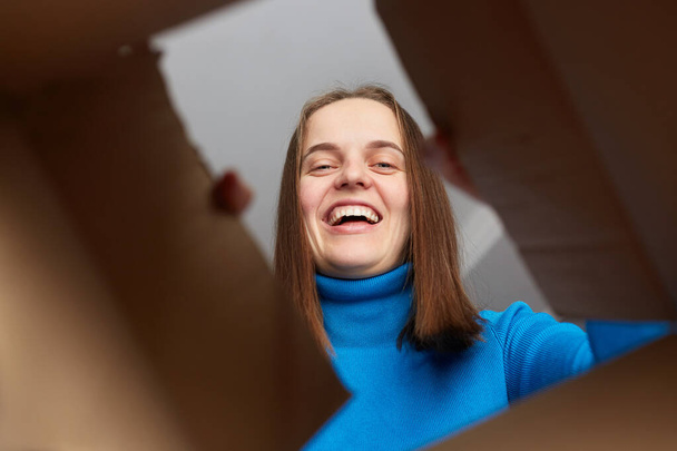 Smiling joyful happy winsome woman wearing blue turtleneck opening cardboard box, looking inside parcel with overjoyed expression, rejoicing her relocation, unpacking her stuff. - Photo, Image