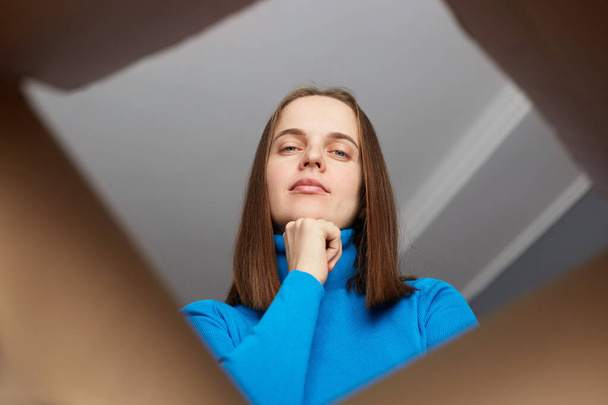 Pensive calm beautiful woman with brown hair wearing blue turtleneck opening cardboard box, looking inside parcel with thoughtful look, holding her chin. - Foto, immagini