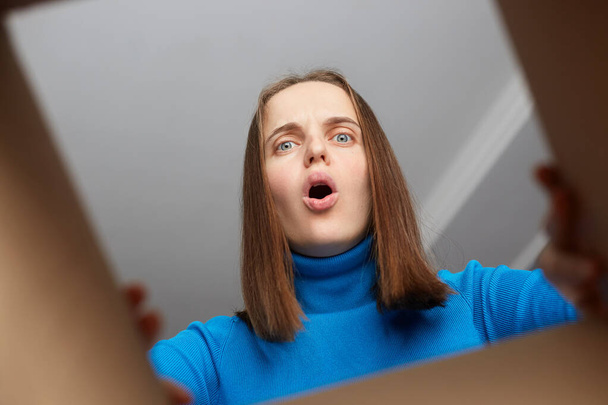 Shocked disappointed woman customer wearing blue turtleneck opening cardboard box with broken or wrong order, looking inside parcel with widely open mouth and big eyes. - Foto, Imagen