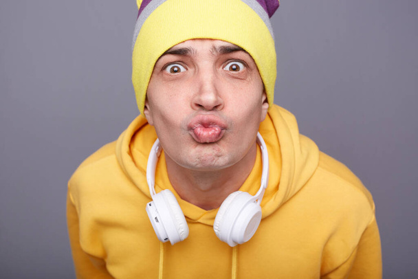 Closeup photo of romantic funny young adult man wearing beanie hat and yellow casual hoodie standing isolated over gray background, keeps lips pout, sending air kisses. - Foto, Imagen