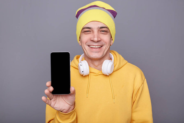 Smiling kind friendly man in beanie hat and yellow casual hoodie, holding mobile phone with blank screen, standing isolated over gray background, showing phone with empty display for advertisement. - Photo, Image