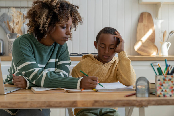 Εστιασμένη μητέρα σημύδας εξηγώντας δύσκολη σχολική εργασία στο προσεκτικό γιο για αποτελεσματική μάθηση. African American προσωπική δάσκαλος γυναίκα και μικρό σχολιαρόπαιδο κάθονται στο γραφείο στο σπίτι μελέτη κουζίνα μαζί - Φωτογραφία, εικόνα