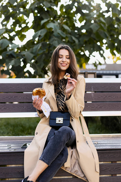 Menina bonita está sentada em um banco na rua, comendo um croissant fresco e delicioso. Uma jovem rompe um pedaço de croissant. - Foto, Imagem