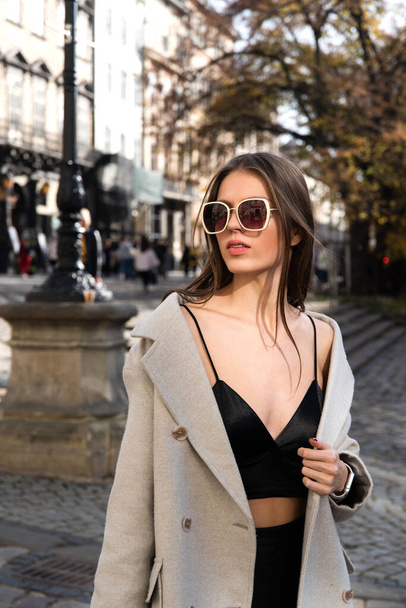 charming brunette posing on the street in spring, wearing a stylish beige coat, a top with razors and sunglasses - Photo, Image