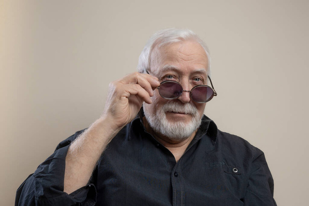 Харизматичный пожилой седой мужчина в солнцезащитных очках и серой рубашке позирует перед камерой в студии, смотрит с улыбкой, крупным планом, светлым фоном - Фото, изображение