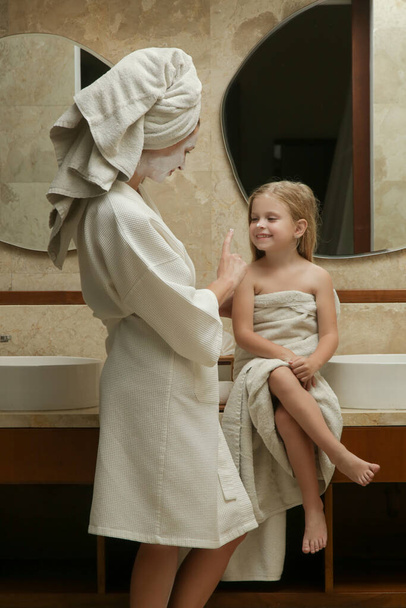 Porträt einer Mutter mit ihrer kleinen, in Handtücher gehüllten Tochter, die im Badezimmer Spaß bei der Hautpflege und guter Familienzeit hat - Foto, Bild