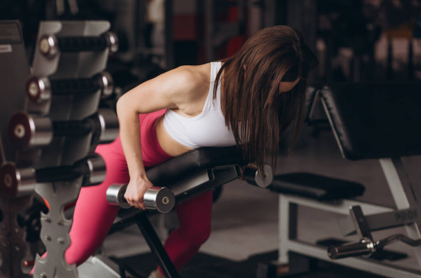 Een sterke bodybuilder leunt op een bank in een sportschool en doet workouts voor ruggen met halters. - Foto, afbeelding