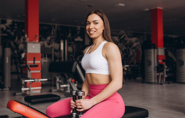 fitness girl with dumbbells posing on a bench in the gym - Photo, Image
