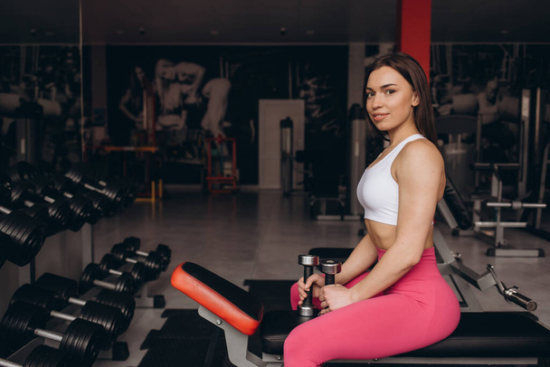 Uśmiechnięta kobieta sport trening biceps z hantlem w dłoni podczas siedzenia na ławce w siłowni - Zdjęcie, obraz