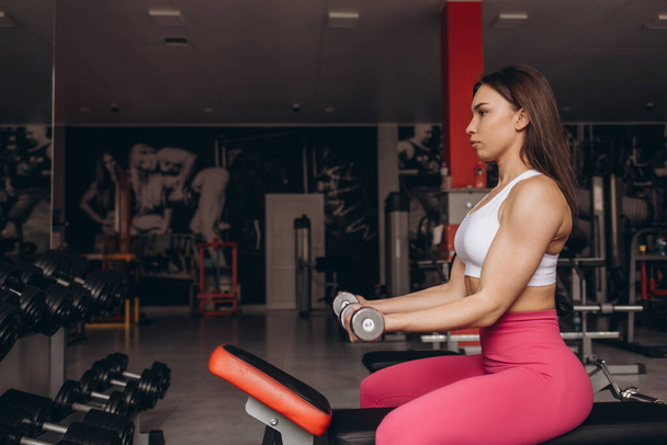 Usmívající se sportovní žena trénink biceps s činkou v ruce, zatímco sedí na lavičce v posilovně - Fotografie, Obrázek
