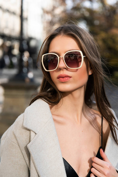 İlkbaharda sokakta poz veren çekici bir esmer, şık bir bej ceket, ustura ve güneş gözlüklü bir bluz giyiyor. - Fotoğraf, Görsel