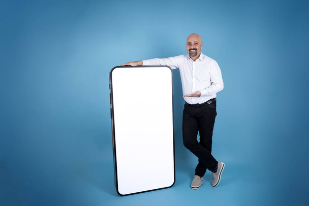 Mobile app advertisement concept, happy man leaning full body length of big cell phone. Modern smartphone mock up idea. Standing on blue studio background. White empty screen, display. Copy space. - Fotografie, Obrázek