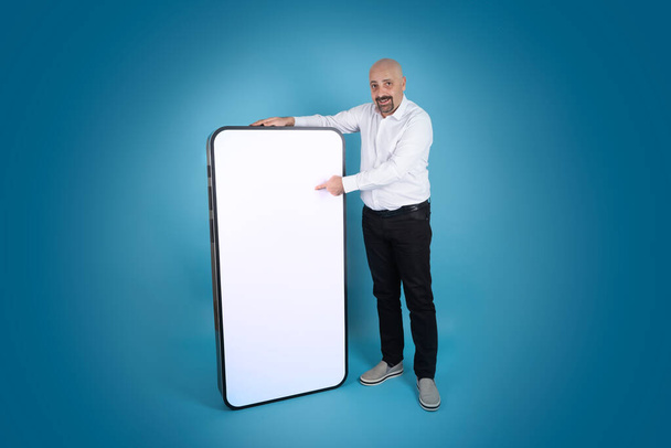 Big cell phone mock up, caucasian man standing by big cell phone mock up. Pointing, touching white blank screen of smartphone. Blue studio background with copy space. Full length body shot. - Photo, Image
