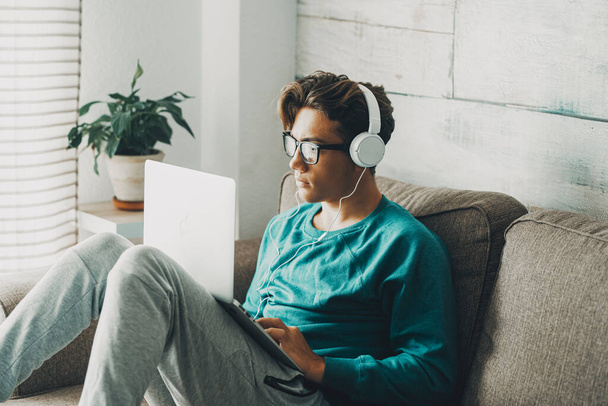 Χαλαρός νεαρός έφηβος που χρησιμοποιεί φορητό υπολογιστή κάθεται στον καναπέ στο σπίτι για παιχνίδι ή μελέτη. Φορώντας ακουστικά. Ένας έφηβος ακούει μουσική σε εσωτερικούς χώρους χαλαρώστε δραστηριότητες αναψυχής και μόνο. Απογευματινή ώρα μόνη - Φωτογραφία, εικόνα