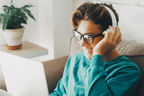 Boldog fiatal diák tinédzser fiú zenét hallgatni vagy leckét fülhallgató és a laptop használata a kanapén délután beltéri szabadidős tevékenység otthon egyedül. Vidám srác jóképű videó hívás iskolai osztály - Fotó, kép