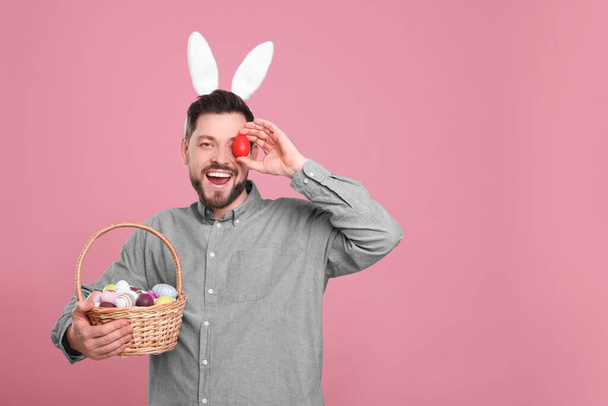 Hombre feliz con diadema de orejas de conejo sosteniendo canasta de mimbre con huevos de Pascua pintados sobre fondo rosa. Espacio para texto - Foto, Imagen