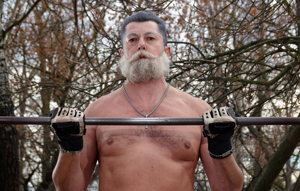 Kiev, Ucrania 11 de noviembre de 2021: Un abuelo joven bombea los músculos con una barra y pesas en una cadena - Foto, Imagen