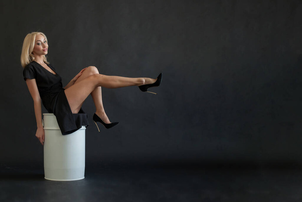 Beautiful surprised shocked blonde woman in black dress sitting white barrel, raise leg up on black studio wall background - Photo, Image
