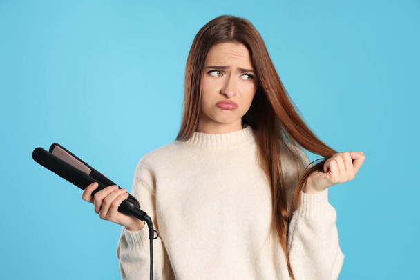 Upset young woman with flattening iron on light blue background. Hair damage - 写真・画像