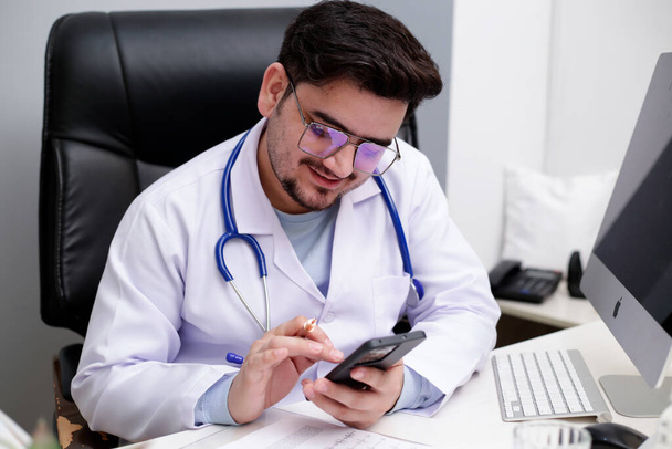 Un médico joven está sentado en la clínica y viendo algo en el teléfono móvil. - Foto, Imagen