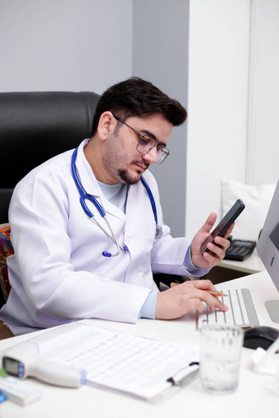 Een jonge arts zit in de kliniek met een mobiele hand en typt iets op het toetsenbord.. - Foto, afbeelding