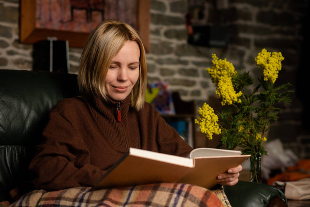 A middle-aged woman, forty years old, is reading a book on the couch. Woman in warm clothes covered with a blanket. Dark key photography. - Foto, Imagem