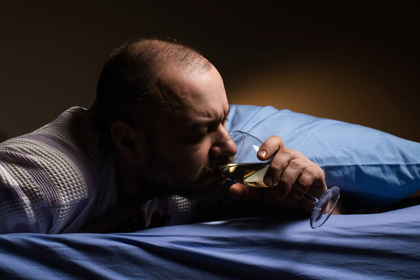 A man alone with a glass of wine in bed. Alcoholism, loneliness. Drink alcohol to sleep. A dark room. - Photo, Image