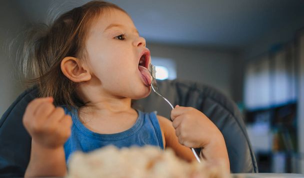 Una niña pequeña sentada en una trona y comiendo comida de un plato con un tenedor. Utensilios para la alimentación de bebés - Foto, Imagen