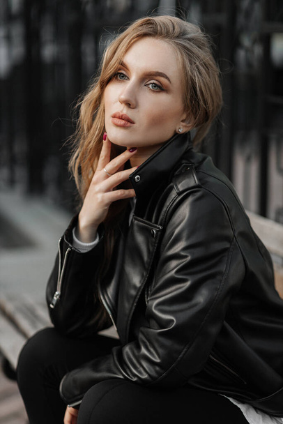 Retrato de cerca de una hermosa joven rubia sonriente en una chaqueta de cuero negro en el fondo de una calle de la ciudad - Foto, Imagen