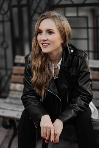 close-up portret van een mooie glimlachende jonge blonde vrouw in een zwart lederen jas op de een stad straat achtergrond - Foto, afbeelding