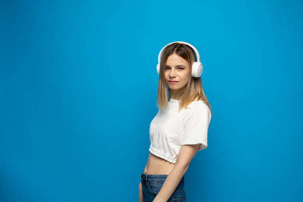 Cheerful young pretty girl smiling while listening music in headphones and dancing on blue background - Foto, immagini