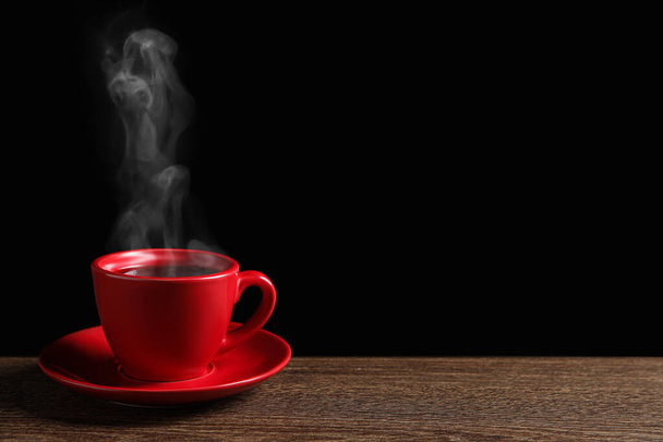 Red cup with hot steaming coffee on wooden table against black background, space for text - Fotó, kép