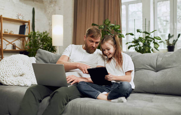 Father giving attention to his little daughter playing on tablet. Man working on laptop at home in living room. Concept of fatherhood, childhood, family, freelance job, home office - 写真・画像