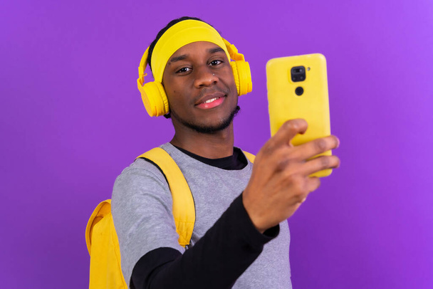 Black ethnic man with backpack and yellow headphones on a purple background, student concept, concept listening to music online - Фото, изображение