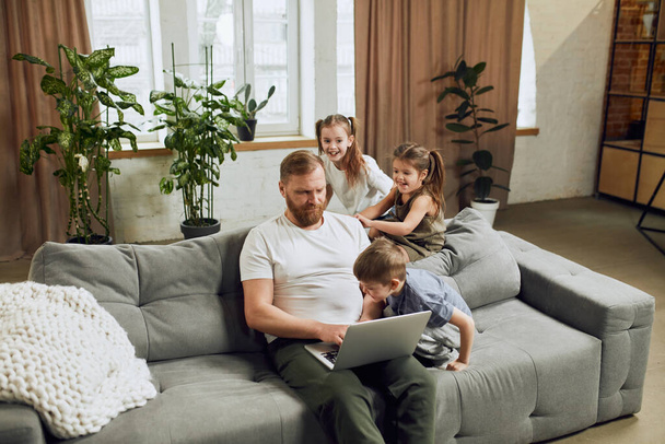 Mature man, father sitting on couch at home and working remotely on laptop with three little kids playing around him and making noise. Fatherhood, childhood, family, freelance job, home office concept - Foto, afbeelding