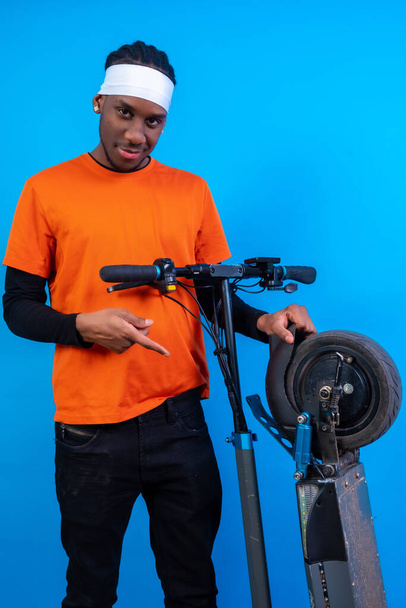 Portrait of a black ethnic man in orange clothes on a blue background, with an electric skateboard smiling - Φωτογραφία, εικόνα