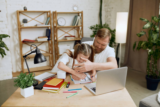 Mature man, father sitting at table and working on laptop remotely at home with little girl drawing, playing with him. Concept of fatherhood, childhood, family, freelance job, home office - 写真・画像