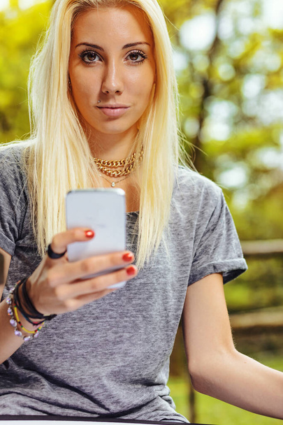 Beautiful blond girl in a park, has long hair and cell phone in hand. It could be summer or fall, but she is wearing a short-sleeved gray T-shirt. - Photo, Image